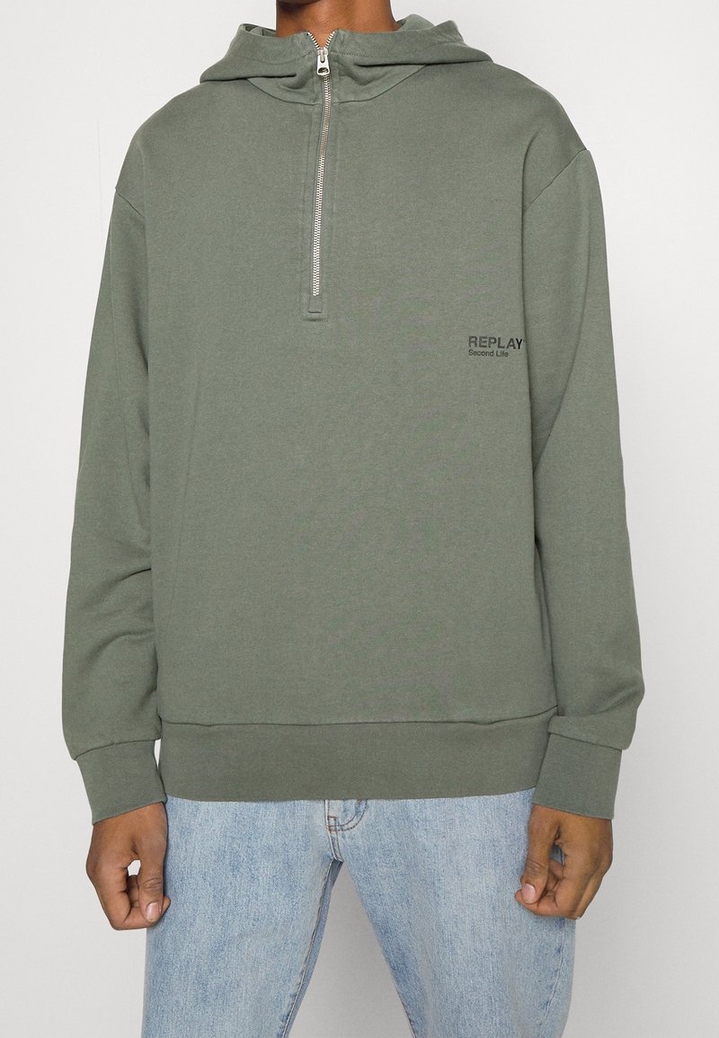 Sudadera con cremallera en verde oliva, con un bolsillo tipo canguro y puños de canalé. Presenta el logotipo "REPLAY" en negro en la parte izquierda del pecho. Tejido suave.