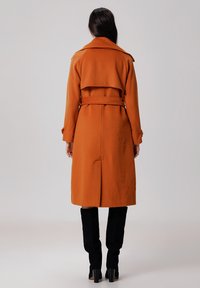 Long manteau orange avec une taille ceinturée, un large col et une fente à l'arrière. Un tissu texturé et un design ajusté mettent en valeur sa silhouette structurée.