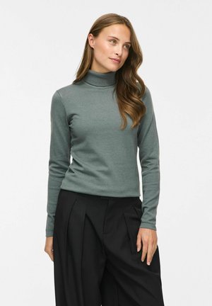 Femme aux longs cheveux portant un pull à col roulé vert et un pantalon noir ample, debout devant un fond clair uni.