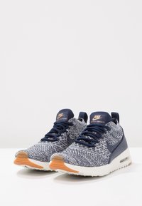 Sneakers Nike Air Max Thea Ultra mají pletený svršek v námořnické modři a bílé barvě, s černými tkaničkami, gumovou podrážkou a béžovým akcentem na špičce.