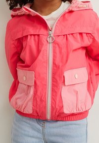 Bambino che indossa una giacca con cappuccio di un vivace colore rosa, con tasche frontali e zip, abbinata a jeans di denim azzurro chiaro.