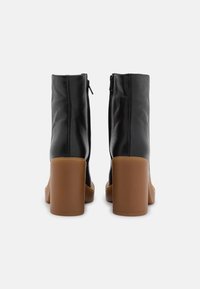 Botas de cuero negras hasta el tobillo con cierre de cremallera, con un tacón grueso marrón y una textura suave. Altura del tacón aproximadamente 10 cm.