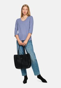 Schwarze Ledertasche mit langen Trägern und einem vorderen Klappdeckel, kombiniert mit einem helllila Oberteil und blauen Jeans. Einfarbig und klassisches Design.