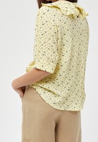 Blusa gialla texturizzata con colletto a balze e piccoli motivi floreali, abbinata a pantaloni beige. Presenta una vestibilità rilassata e maniche corte.