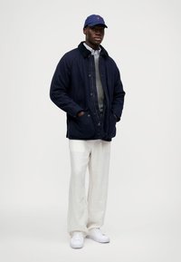 Casaco de mistura de lã azul-marinho com gola, suéter cinza por baixo, calças brancas de linho e ténis brancos. Acessórios incluem um boné azul.