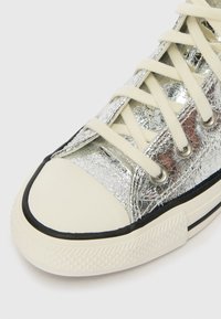 Sapatilha metálica prateada com um upper texturizado, atacadores creme e uma ponteira de borracha branca. linha de destaque preta ao longo da sola.