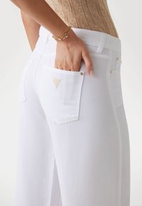 Jeans in denim bianco con vita alta, dotati di una grande tasca posteriore, dettagli in oro sottili e un logo di marca triangolare sulla tasca.