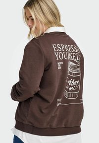 Blondýnka ve hnědém svetru s bílou košilí, vzadu na svetru je grafika hrnku kávy a text "Espresso Yourself, Coffee to go."