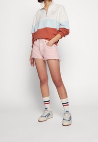Persona que lleva un sudadera con bloques de color, pantalones cortos de mezclilla rosa claro, calcetines blancos con rayas rojas y azules, y zapatillas blancas con detalles azules.