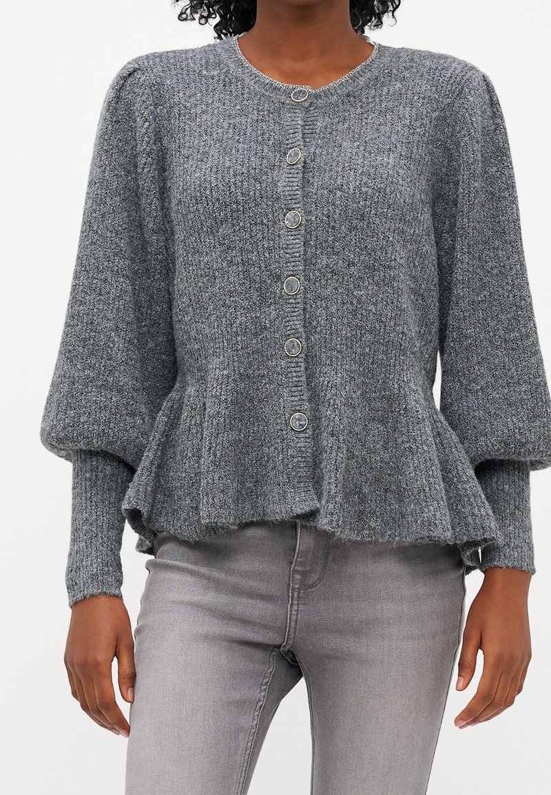 Femme portant un cardigan en maille gris boutonné avec un ourlet évasé et des manches trois-quarts bouffantes, associé à un jean gris clair.