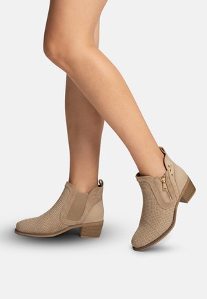 Beige geperforeerde enkellaarzen met ritssluitingen aan de zijkant en blokhakken, gedragen op gekruiste benen tegen een effen achtergrond.