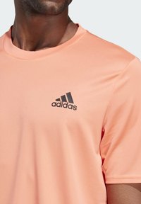 Camiseta deportiva de color rosa claro fabricada con tejido texturizado, con un logo negro de Adidas en el lado izquierdo del pecho y un escote redondo.
