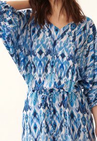 Modrá vzorovaná šaty s výstřihem do V a krátkými rukávy, s detailem vázání v pase. Tkanina je lehká a splývavá.