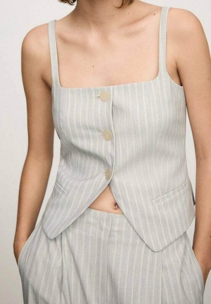 Femme portant un gilet sans manches gris clair à fines rayures avec trois boutons et un pantalon taille haute assorti, mains dans les poches.