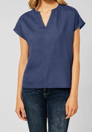 Femme portant un chemisier bleu marine à manches courtes et col en V associé à un jean bleu foncé sur un fond uni.