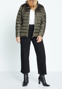 Chaqueta acolchada en verde oliva, diseño acolchado, cuello alto, cierre con cremallera. Combinada con unos vaqueros cortos negros y botas de tacón negras.