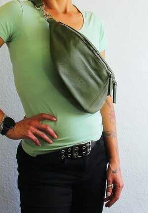 Olive green leather sling bag with a textured surface, zip closure, and adjustable shoulder strap, worn against a light green shirt.