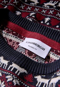 Stickad tröja med en marinblå bas och ett mönster av krämfärgade renar och röda detaljer. Etiketten säger "LINDBERGH" och "AVSLAPPNAD PASSFORM."