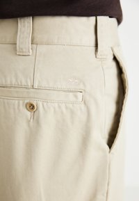 Beige cotton shorts with front and back pockets, featuring a visible button and a small embroidered logo above the rear pocket.
