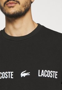 Czarna bawełniana koszulka z okrągłym dekoltem; na przodzie znajduje się biały napis "LACOSTE" oraz logo krokodyla. Miękka tekstura, luźny krój.