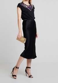 Frau in schwarzem Oberteil mit buntem Perlenkragen, schwarzem satin Midi-Rock, schwarzen Ankle-Strap-Absätzen und einer goldenen Clutch.