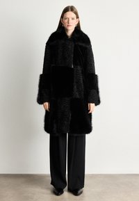 Melnas kažokādas mētelis ar apdarēm, kas sastāv no dažādām faktūrām un garumiem. Augsts apkakle un plašas piedurknes. Uzvilkts virs melniem biksēm.