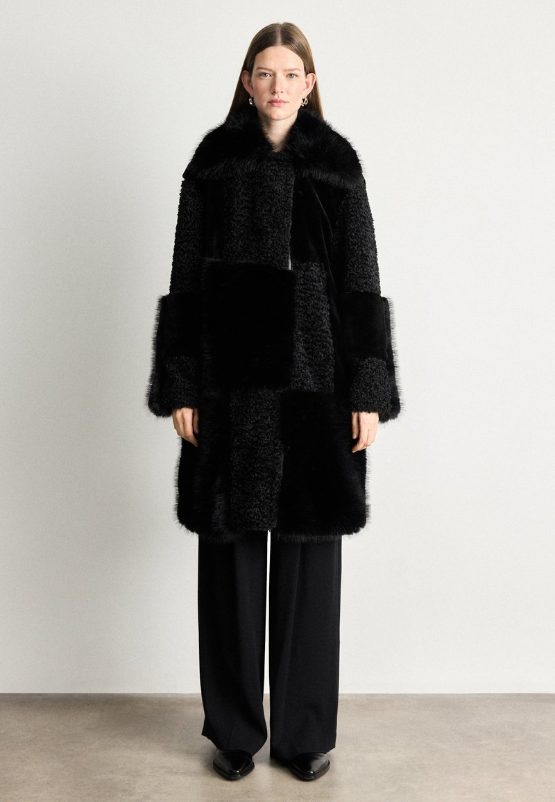 Melnas kažokādas mētelis ar apdarēm, kas sastāv no dažādām faktūrām un garumiem. Augsts apkakle un plašas piedurknes. Uzvilkts virs melniem biksēm.