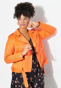 Veste de moto en daim orange avec des poches zippées et un détail de ceinture, superposée sur une robe noire à fleurs, tenue par une personne.