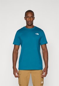 Teal crew-neck t-shirt lavet af bomuld med korteærmer og et hvidt logo på venstre bryst, designet til komfort og udendørs aktiviteter.
