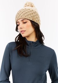 Vrouw die een beige gebreide beanie met pompon en een donkerblauwe rits-top draagt, terwijl ze omlaag kijkt tegen een witte achtergrond.