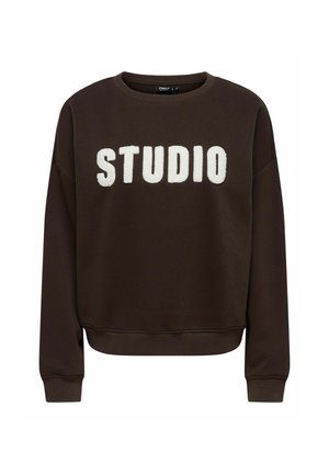 Bruine sweatshirt met een ronde halslijn en geribbelde manchetten, met grote, witte pluizige letters die "STUDIO" op de voorkant spellen.