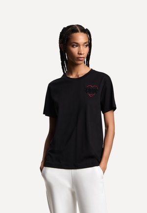 Donna con capelli intrecciati, indossa una maglietta nera con una grafica a cuore rosso e pantaloni bianchi, in piedi con le mani nelle tasche, guarda di lato.