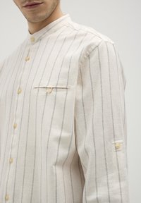 Camicia button-up beige chiaro con sottili righe nere verticali, dotata di colletto alto e taschino sul petto, realizzata in un tessuto testurizzato.