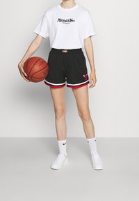 Henkilö, joka käyttää valkoista Mitchell & Ness -t-paitaa ja mustia Chicago Bulls -koripalloshortseja, pitää oranssia koripalloa vasemmassa kädessään.
