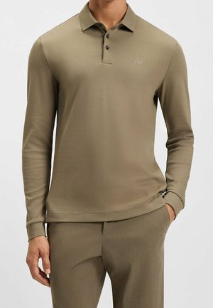 Mann trägt ein langärmliges olivgrünes Polohemd mit drei schwarzen Knöpfen und passende olivgrüne Hose.