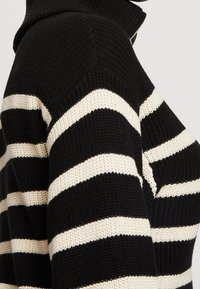 Schwarzer gestrickter Pullover mit dicken horizontalen cremefarbenen Streifen und einem sichtbaren Reißverschluss am hohen Kragen auf dem Oberkörper einer Person.