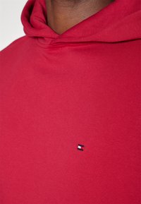 Acercamiento de una persona que lleva puesto un hoodie rojo con un pequeño logo rectangular bordado en el pecho.