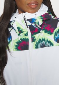 Vit jacka med ett färgglatt blomstertryck i grönt, blått och rött på huvan och övre bröstet, med dragkedja och justerbara snoddar.