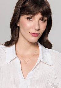 Złoty naszyjnik z delikatnym łańcuszkiem i małym wisiorkiem. Modelka ma na sobie fakturowaną białą bluzkę, co podkreśla naszyjnik na neutralnym tle.