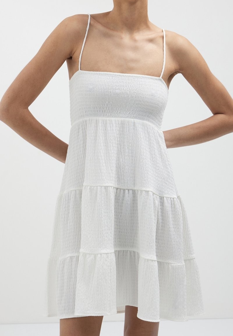 Femme portant une robe blanche sans manches à étages avec de fines bretelles, debout les mains sur les hanches devant un fond clair uni.