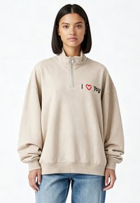 Sweat-shirt beige à fermeture éclair avec col montant, broderie noire et rouge indiquant "I ❤️ YOU", et manches amples.