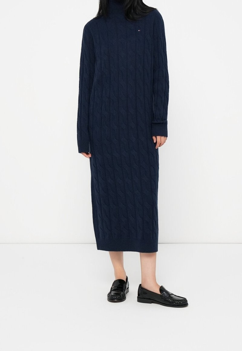 Femme portant une robe midi à col roulé en maille torsadée bleu marine et des mocassins en cuir noir, debout devant un fond blanc.