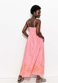 Femme aux cheveux courts et bouclés, portant une robe maxi sans manches rose avec des broderies florales orange et des chaussures à enfiler en imprimé léopard, posant devant un mur blanc.