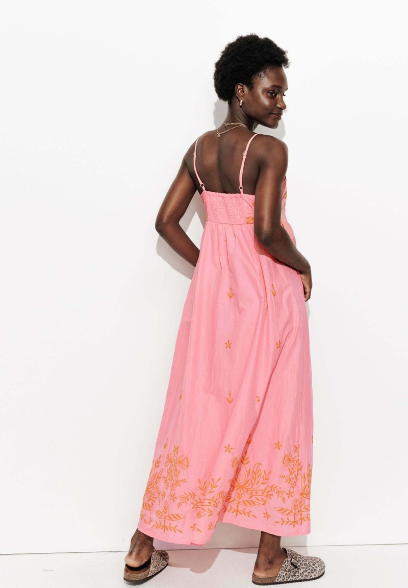 Femme aux cheveux courts et bouclés, portant une robe maxi sans manches rose avec des broderies florales orange et des chaussures à enfiler en imprimé léopard, posant devant un mur blanc.