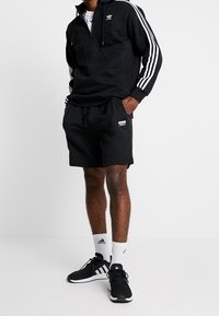 Svart zip-up-hoodie med vita ränder, kombinerad med svarta shorts. Svarta löparskor och vita strumpor med svarta detaljer fullbordar outfiten.