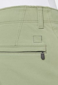 Pantalones de algodón verdes con un bolsillo con cremallera que tiene un tirador negro, detalles de costuras visibles y un acabado de tela texturizado.