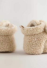 Chaussons en polaire d'un beige doux avec une texture duveteuse, dotés de bords repliés et de petites anses pour un enfilage facile. Agrémentés de tirettes rayées.