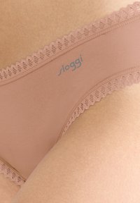 Tan Sloggi tanga ar mežģīņu apdari jostā, kas piedāvā gludu auduma tekstūru un delikātu logotipa detaļu.