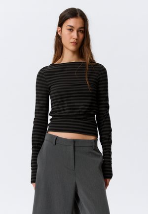 Jeune femme aux longs cheveux bruns portant un haut noir à manches longues rayé et un pantalon gris taille haute à jambes larges, sur un fond uni.