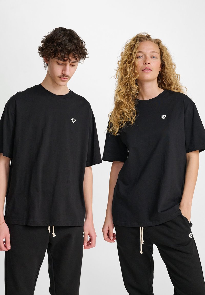 Camisetas de algodón negras con mangas cortas, de corte relajado, cada una con un pequeño logo blanco en el pecho. Llevadas con pantalones negros a juego.